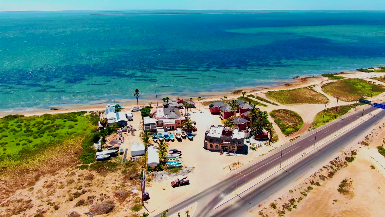 Drone view of coastal buildings, road, dunes, and blue water.