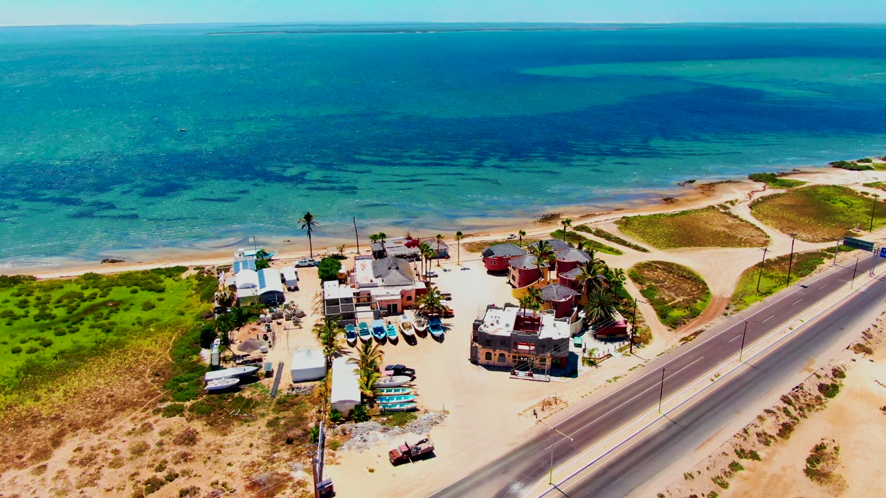 High aerial view of San Carlos, Baja California Sur with the road running beside the sand, beach structures and palms below, green dune patches, blue ocean bands, and no visible people.