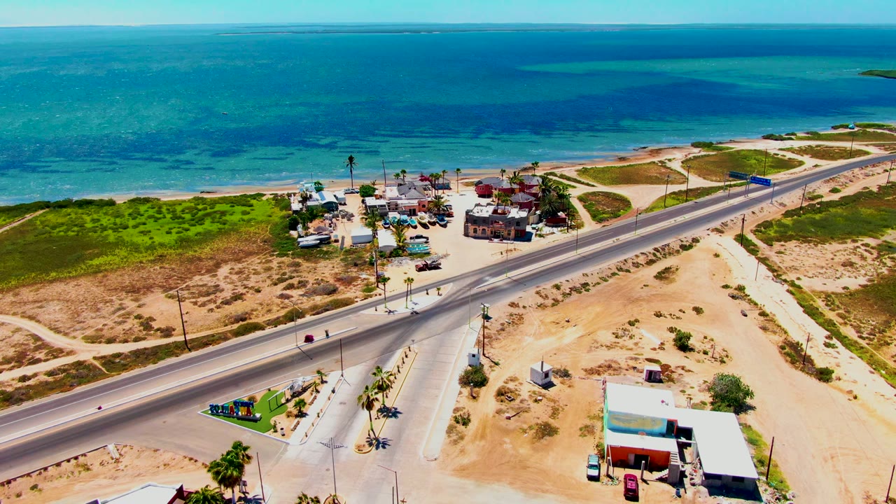 Aerial frame over San Carlos, Baja California Sur, showing a coastal highway, sandy lots, beachside buildings, green scrub, turquoise ocean, and no visible people at the start of the clip.