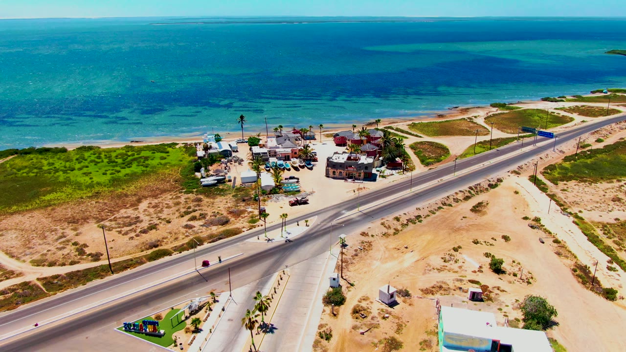 Wide aerial frame of San Carlos, Baja California Sur, showing scattered traffic on a coastal highway, clustered beachside buildings, dry scrubland, turquoise ocean, and no visible people.