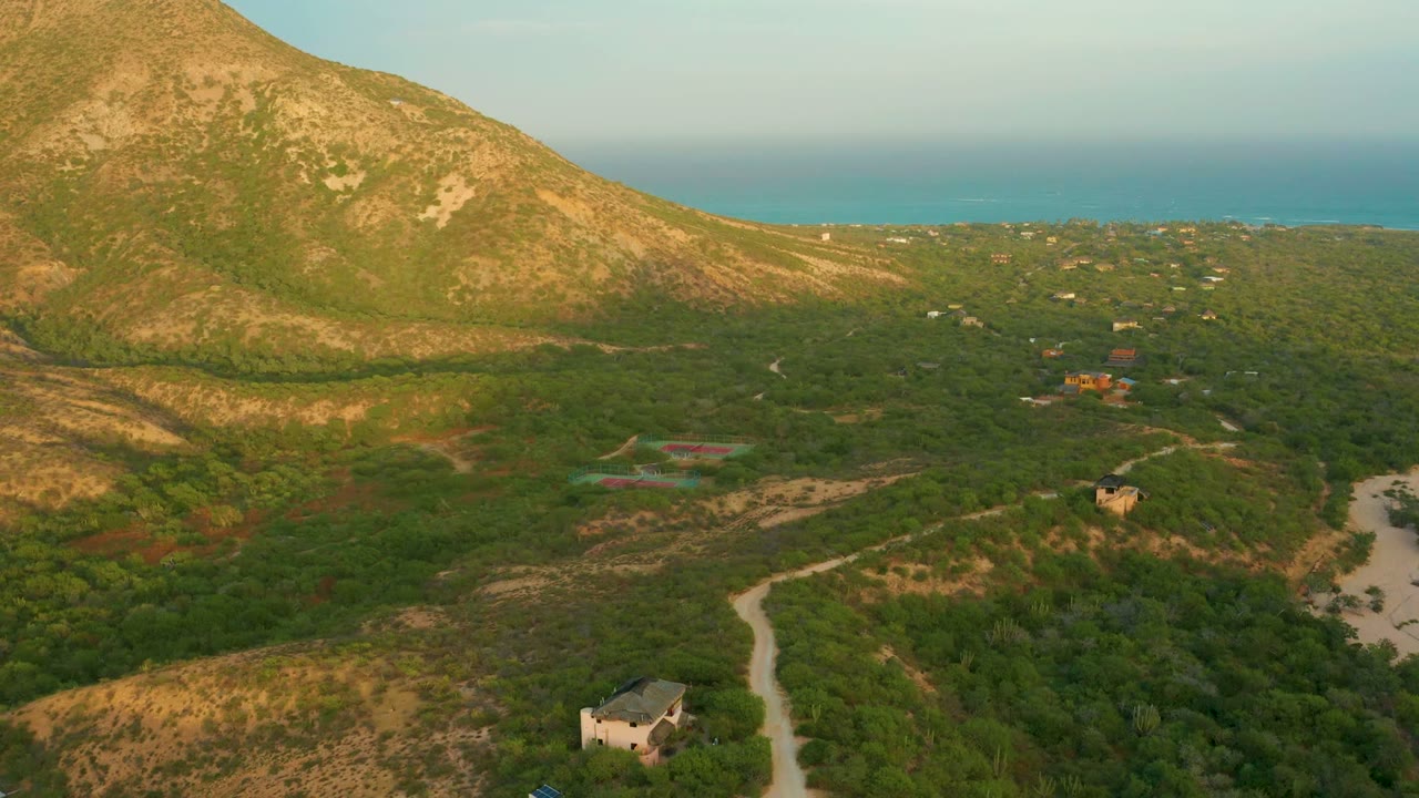 At 0:00, an aerial view in Cabo Pulmo, Baja California Sur shows a pale dirt road through green scrub, small buildings, a sunlit mountain slope, ocean on the horizon, and no visible people.