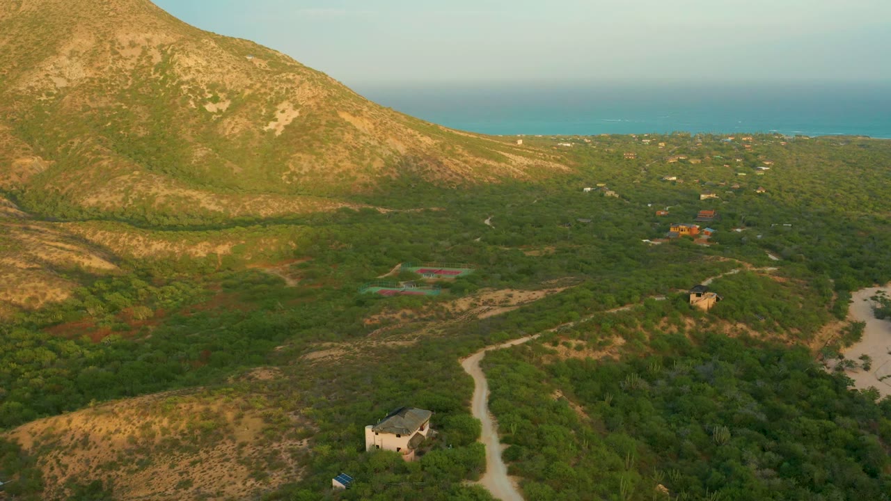 Wide drone view of scrubland, dirt road, buildings, mountain slope, and blue coastline.