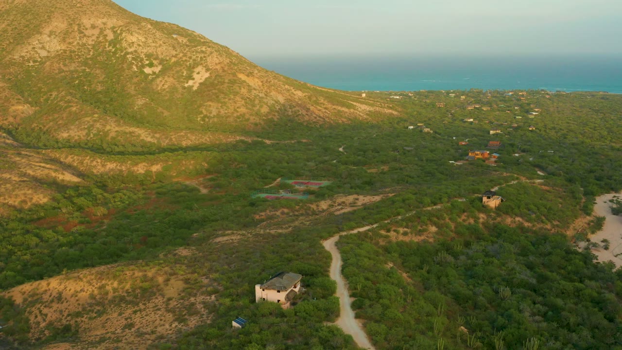At 0:02, the drone view over Cabo Pulmo shows the winding dirt road nearer the lower right, scattered buildings in the greenery, the broad hillside, distant ocean, and no visible people.