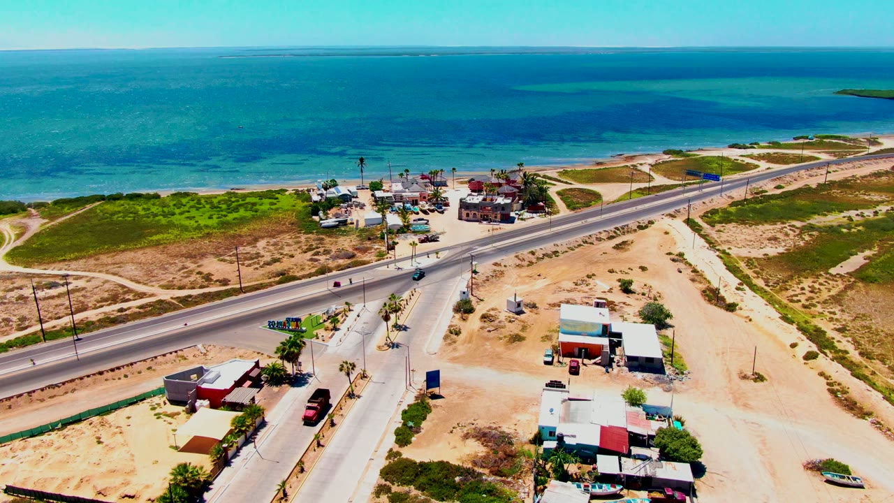 Aerial frame over San Carlos, Baja California Sur, with a paved road, small roadside buildings, sandy lots, green scrub, turquoise ocean, and no visible people.