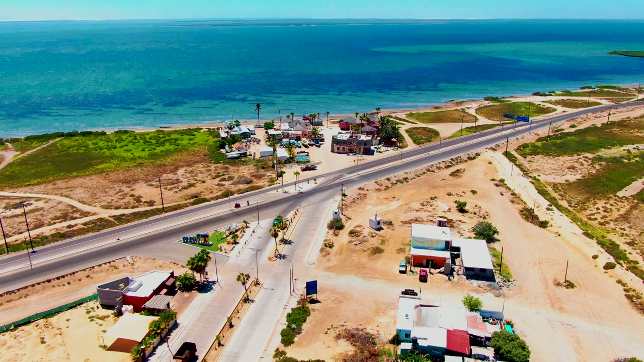 Drone-height view in San Carlos, Baja California Sur, with the coastal road cutting past sandy open land, scattered buildings, roadside poles, turquoise ocean, and no visible people.