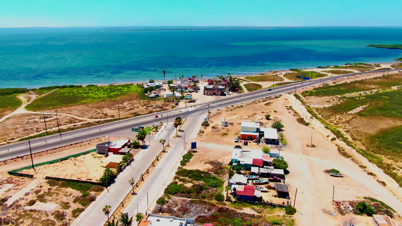 Opening aerial frame over San Carlos, Baja California Sur, showing a paved coastal road, small roadside buildings, sandy terrain, green scrub, calm blue ocean, and no visible people.
