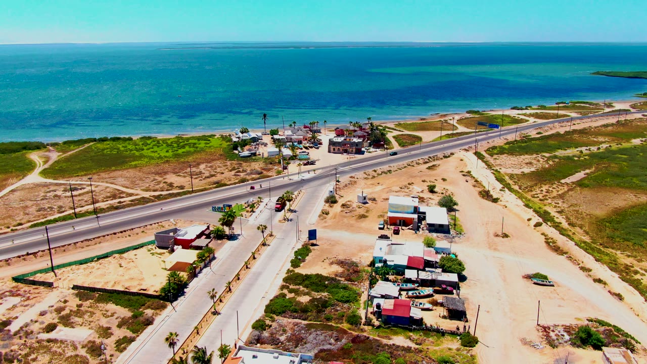 At 0:02, the drone perspective holds above San Carlos, Baja California Sur, with the road running across the frame, clustered low buildings, sandy ground, turquoise water, and no visible people.