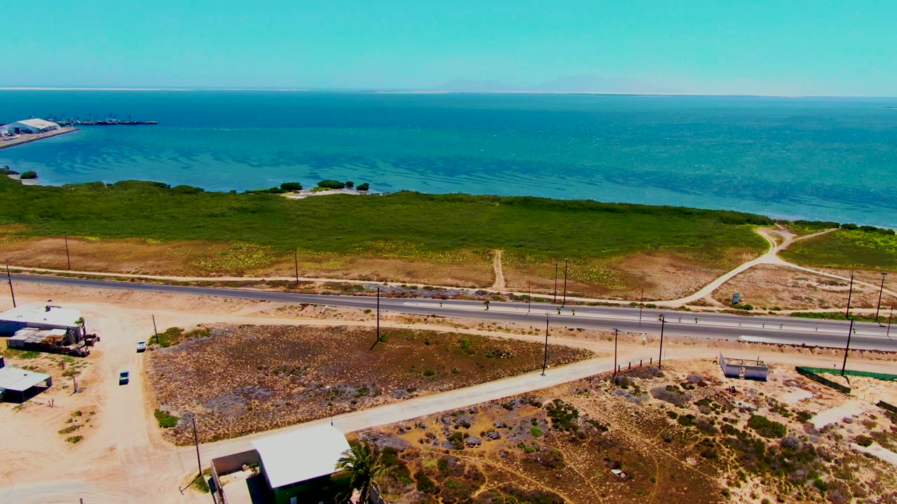 At 0:00, an aerial view in San Carlos, Baja California Sur shows a paved coastal road, dirt tracks, green scrub, dry ground, blue ocean, and no visible people.