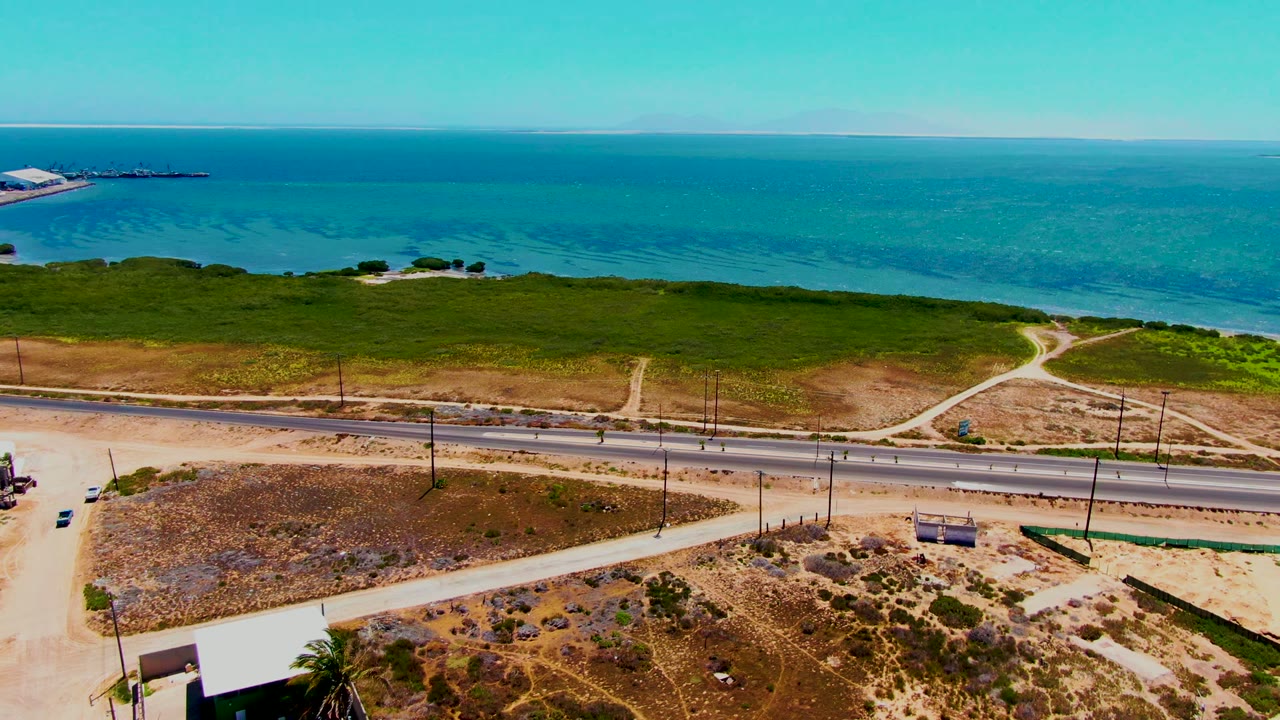 At 0:02, the elevated view in San Carlos shows the highway crossing the lower frame, pale dirt roads cutting toward green coastal vegetation, and no visible people.