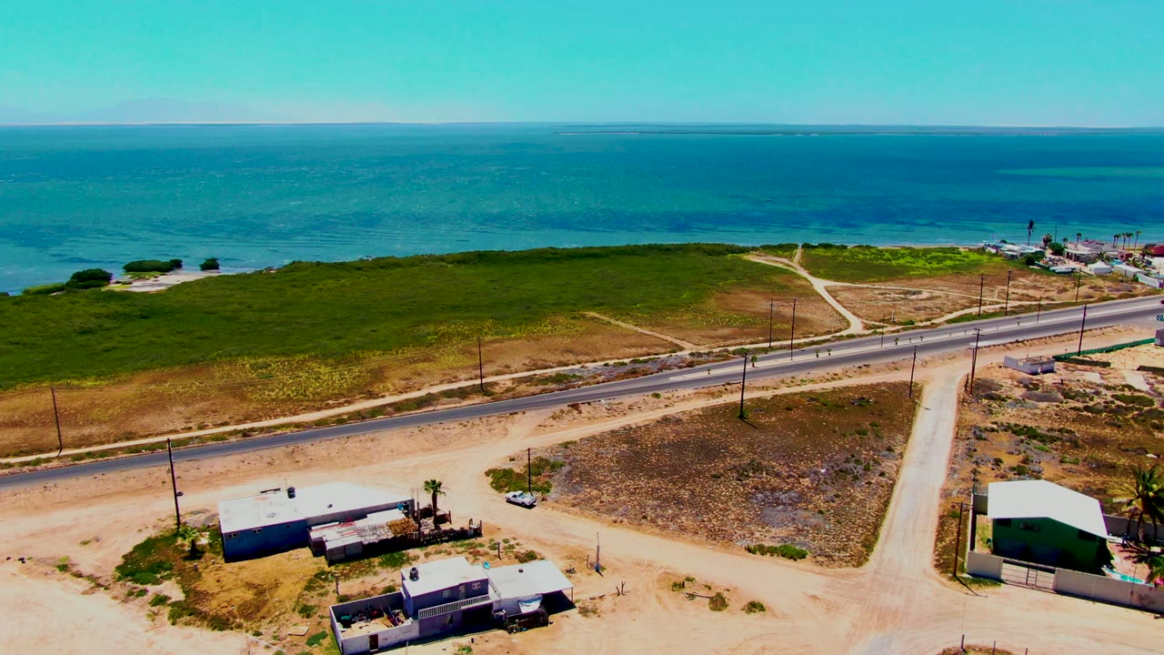 Aerial frame from San Carlos, Baja California Sur showing turquoise ocean, green coastal brush, a paved road, sandy foreground lots, small buildings, utility poles, and no visible people.
