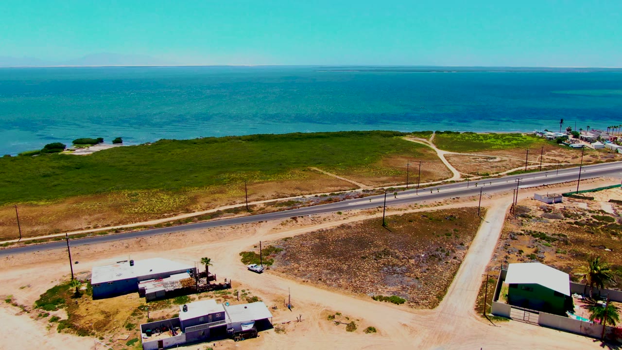 Aerial frame from San Carlos, Baja California Sur with the white car visible on the paved road, ocean across the background, sandy lots in the foreground, small buildings, and no visible people.