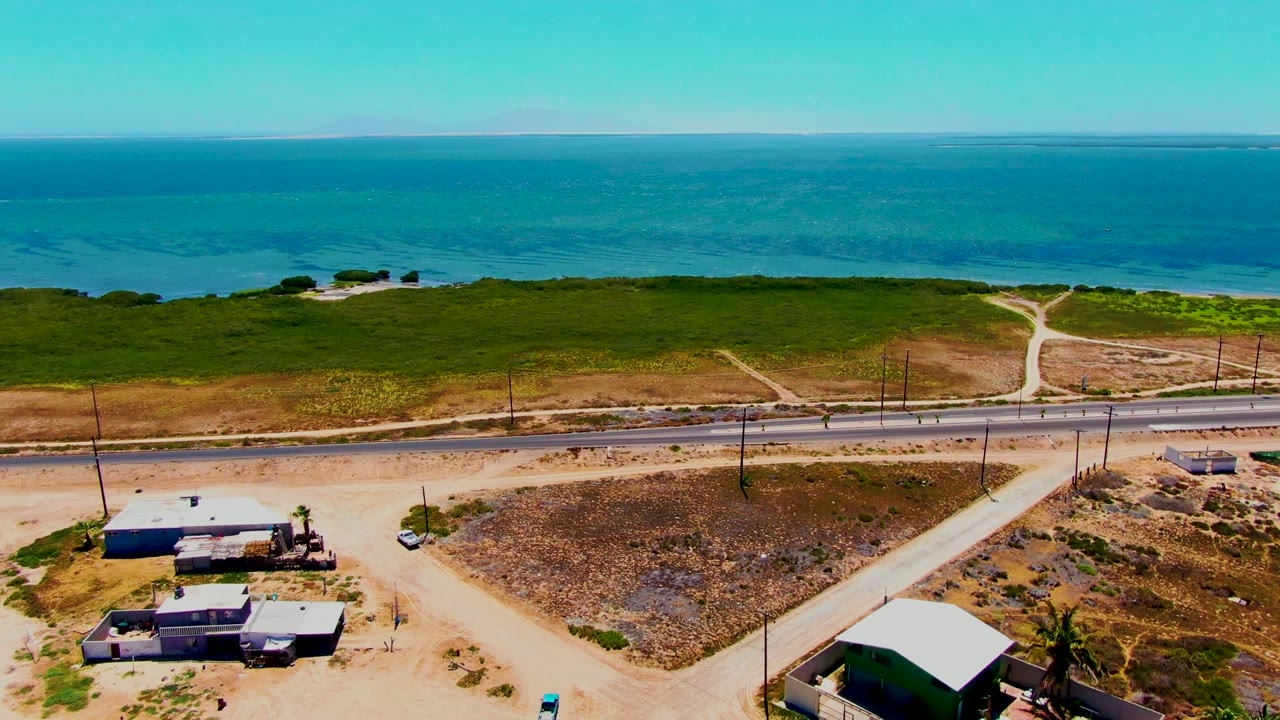 Opening aerial frame in San Carlos, Baja California Sur shows a coastal road below a green shoreline and blue ocean, with small buildings and no visible people.