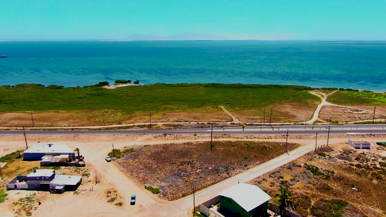 San Carlos aerial view shows dry open lots and a paved road in the foreground, a green strip of coast in the middle, and blue ocean under a clear sky, with no visible people.