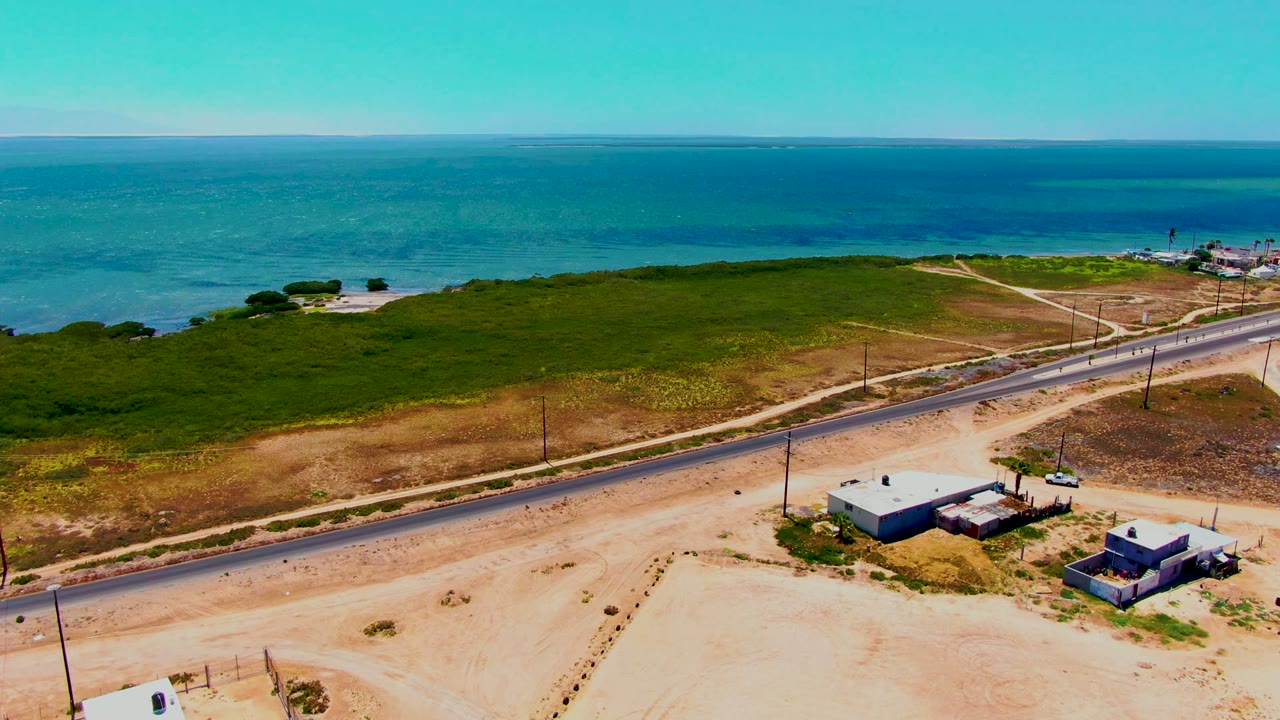Aerial frame in San Carlos, Baja California Sur showing a two-lane road below green shoreline scrub and blue ocean, with sandy foreground lots and no visible people.
