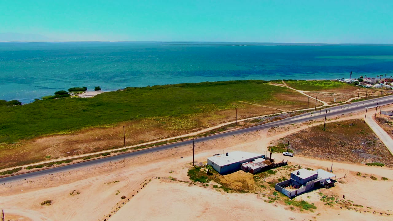 Aerial frame in San Carlos, Baja California Sur showing a road beside sandy access tracks, a cluster of low structures, dense green coastal scrub, blue water, and no visible people.
