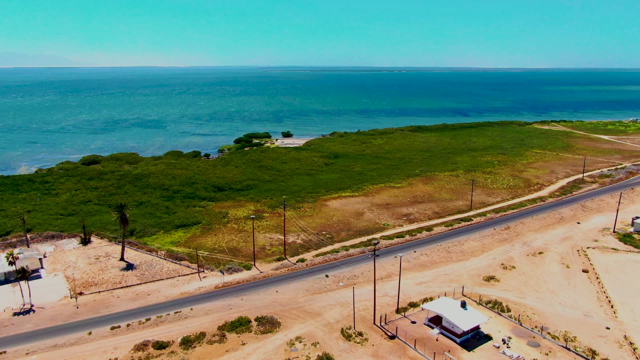 Aerial view in San Carlos, Baja California Sur, showing a coastal road crossing sandy terrain below green vegetation and blue ocean, with no visible people.