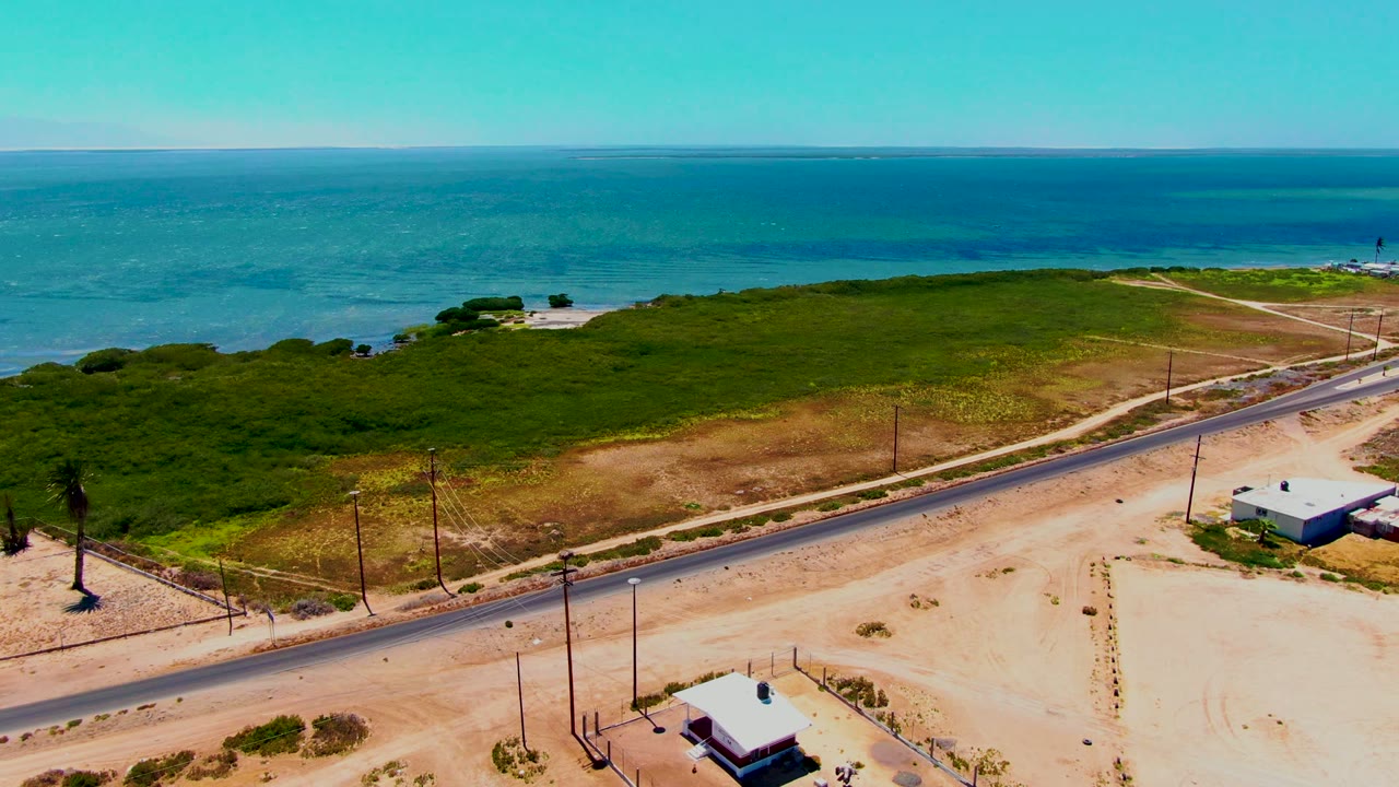 Wide aerial view in San Carlos, Baja California Sur, showing a road along sandy ground, small low buildings near the lower edge, green brush, and the blue ocean horizon, with no visible people.