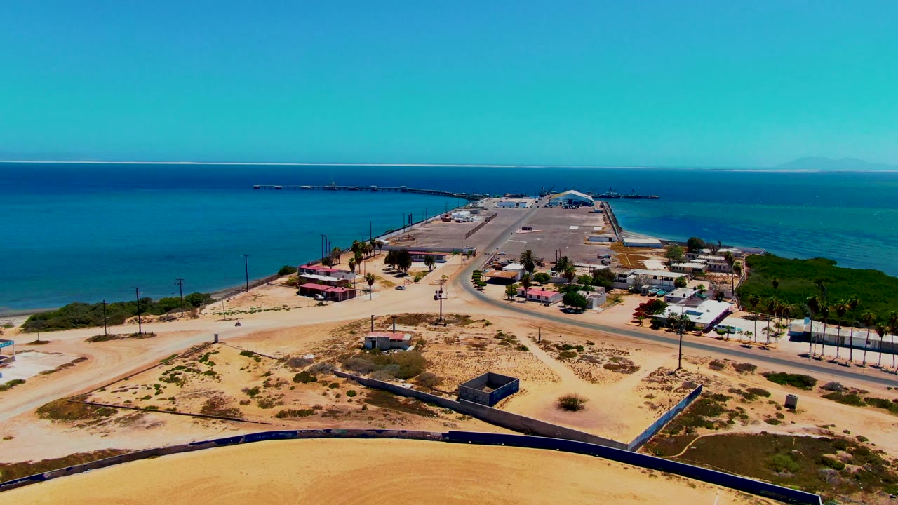 Aerial frame over San Carlos, Baja California Sur showing sandy lots, a paved road, small coastal buildings, a breakwater area, blue sea, distant horizon, and no visible people.
