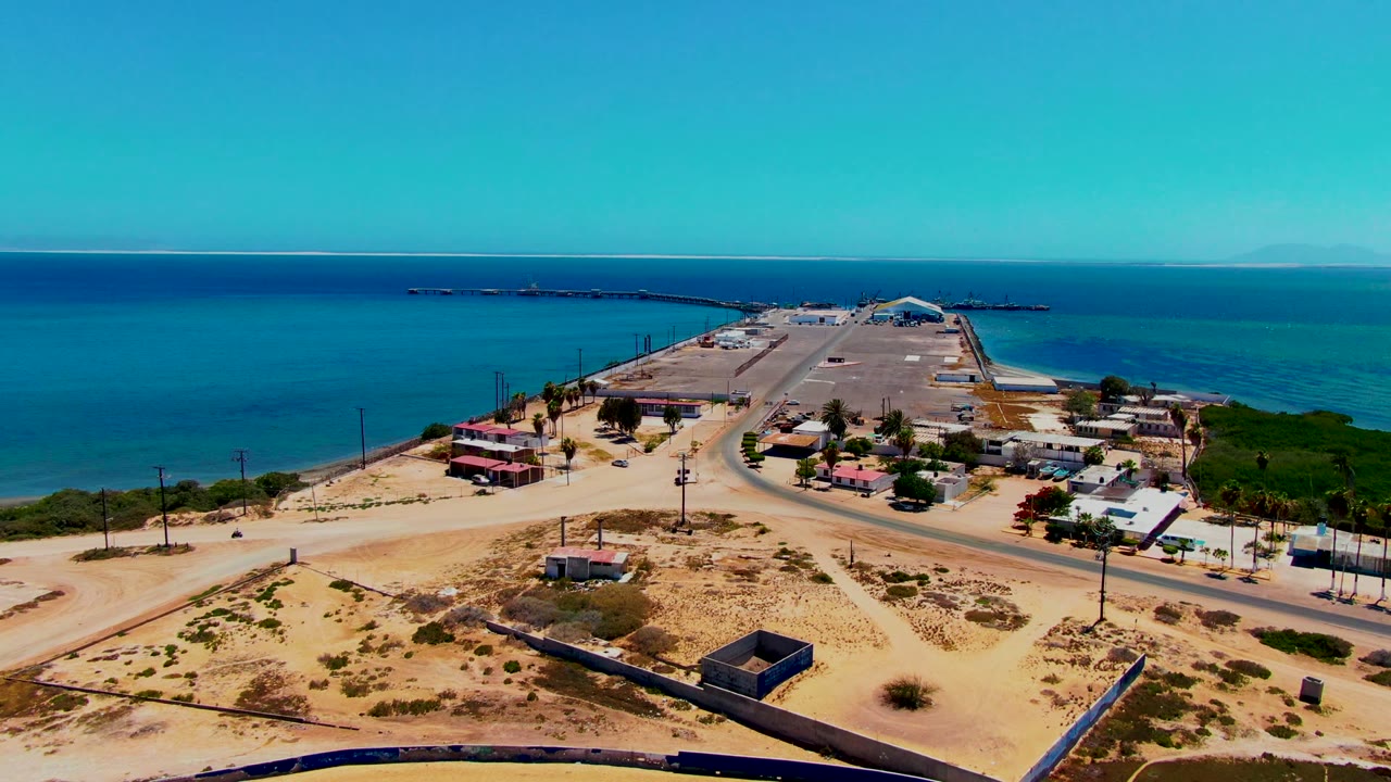 Overhead coastal view of San Carlos, Baja California Sur showing small buildings clustered near a paved road, dry sandy terrain in front, blue ocean beyond, and no visible people.