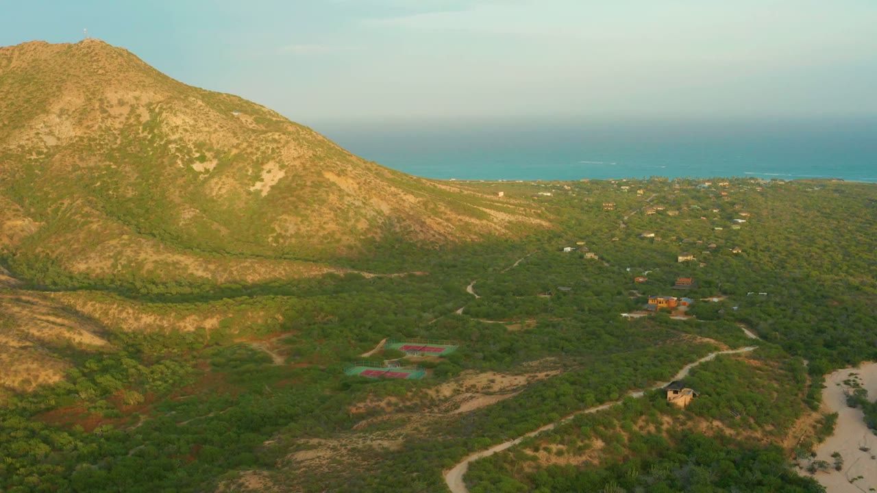 Aerial frame over Cabo Pulmo, Baja California Sur, showing a rocky mountain on the left, dense green valley brush, pale dirt roads, small scattered buildings, a distant blue ocean horizon, and no visible people.