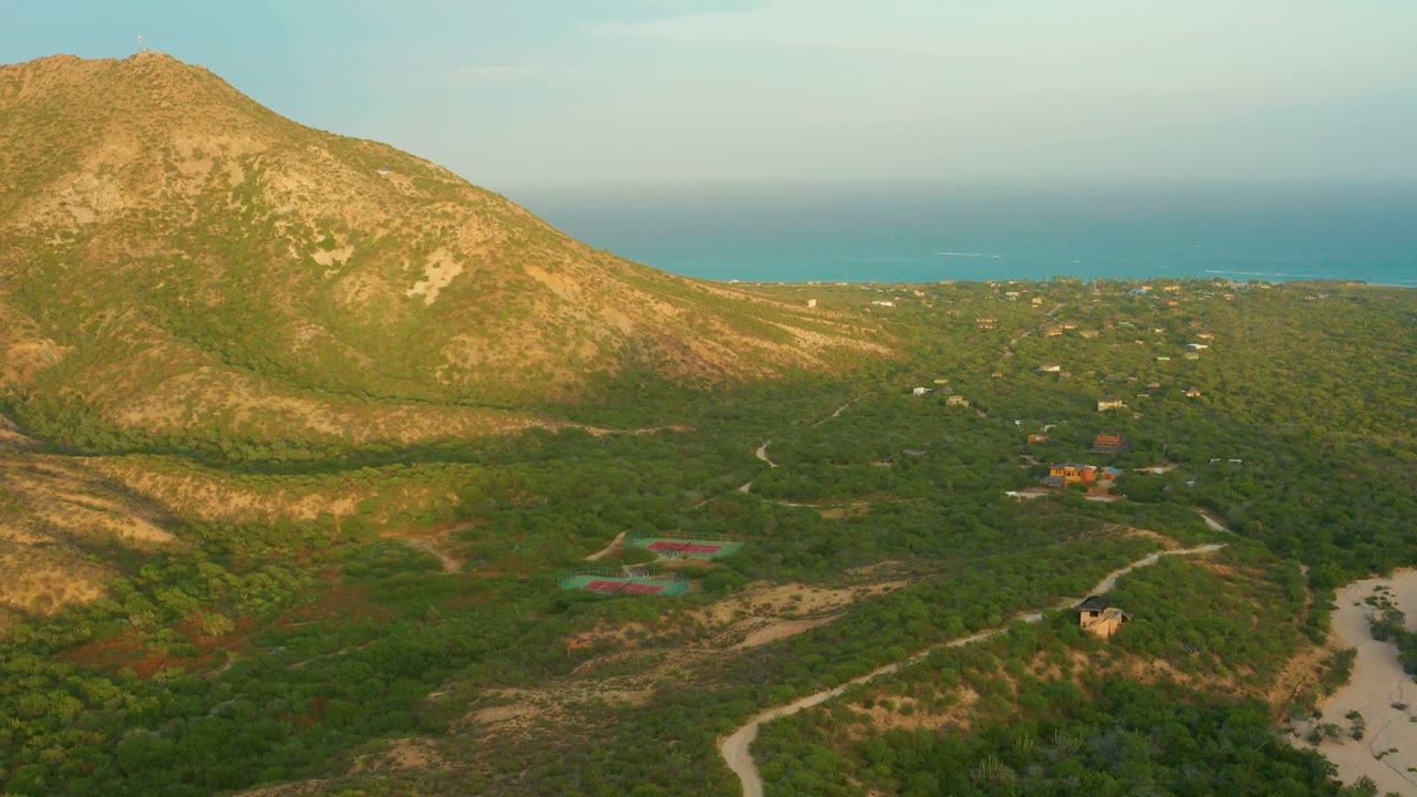 Wide aerial landscape with mountain slope, green scrub valley, roads, buildings, and blue ocean horizon.