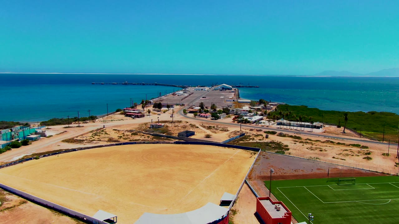 Aerial frame over San Carlos, Baja California Sur, showing a large sandy field, a green sports field, dirt roads, low coastal structures, blue ocean, clear sky, and no visible people.