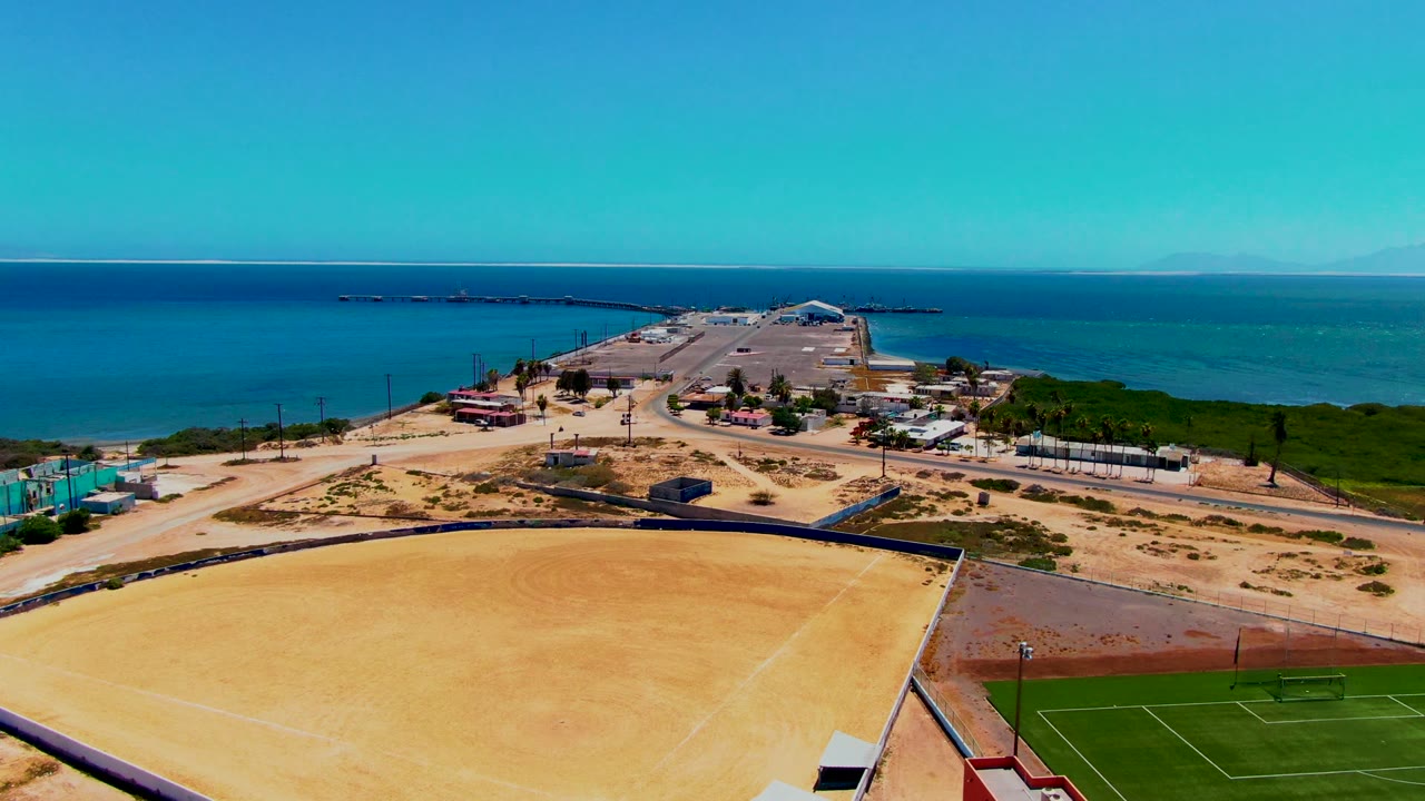 Drone-height view in San Carlos with the sandy field across the lower frame, a green field at right, coastal roads and buildings in the middle distance, ocean beyond, and no visible people.