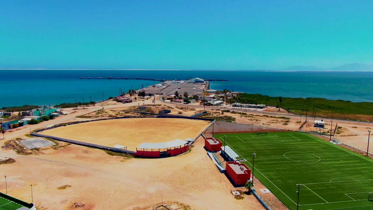 Aerial frame over San Carlos, Baja California Sur showing a green soccer field at right, a sandy arena at left, red structures below, blue ocean beyond, and no visible people.