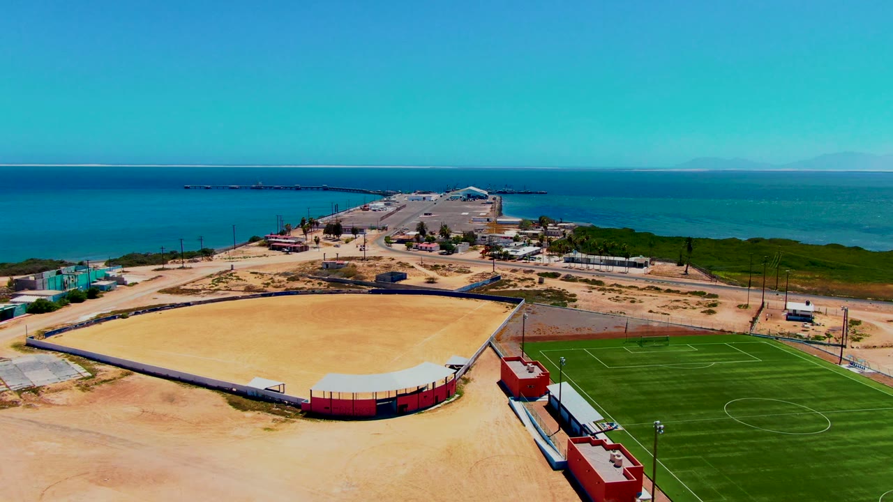 Aerial view over San Carlos showing the sandy arena across the lower center, green turf at the right edge, red buildings near the foreground, ocean on the horizon, and no visible people.