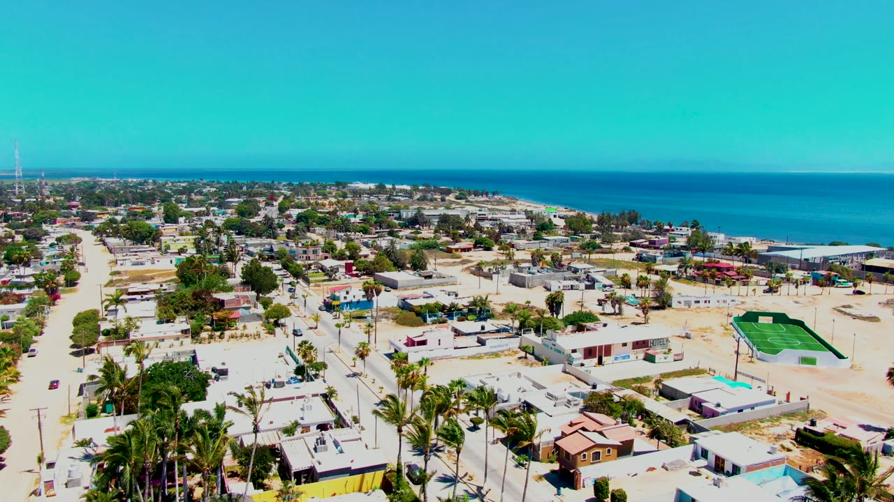 Opening aerial frame over San Carlos, Baja California Sur, showing low-rise homes, sandy streets, palm trees, ocean horizon, blue sky, and no visible people.