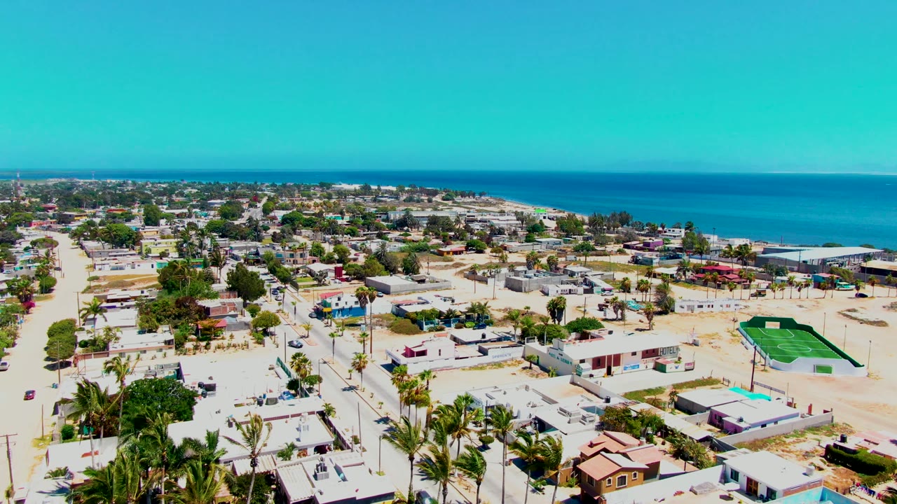 Coastal neighborhood aerial with houses, road grid, palms, and ocean horizon.
