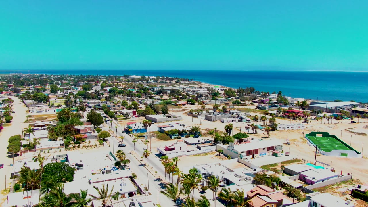 High aerial view of San Carlos town blocks near the coast, with a broad blue ocean band, clear sky, palms, houses, and no visible people.