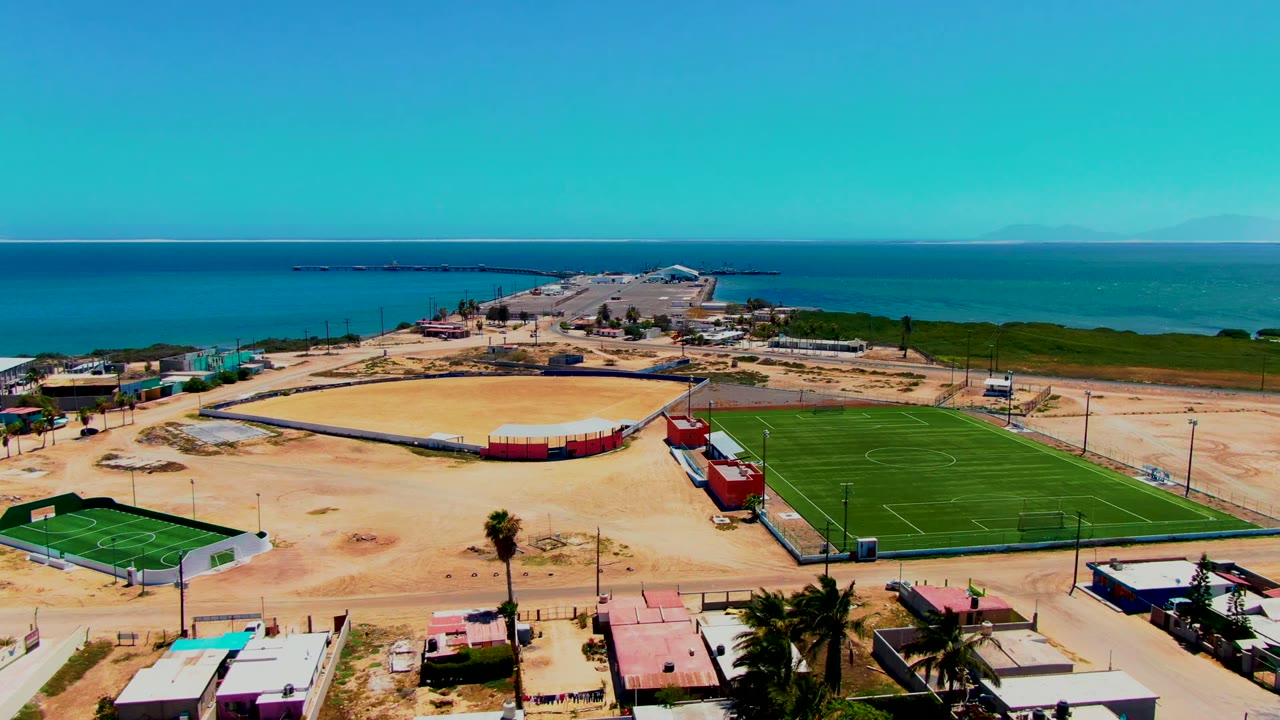 Aerial frame over San Carlos, Baja California Sur, showing green soccer fields, a sandy baseball diamond, palm trees, low buildings, and blue ocean beyond, with no visible people.