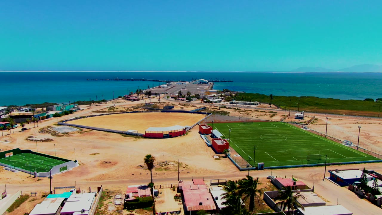 San Carlos, Baja California Sur aerial frame showing the soccer field in the right foreground, the baseball diamond in the center, sandy open ground, low structures, and blue ocean on the horizon, with no visible people.
