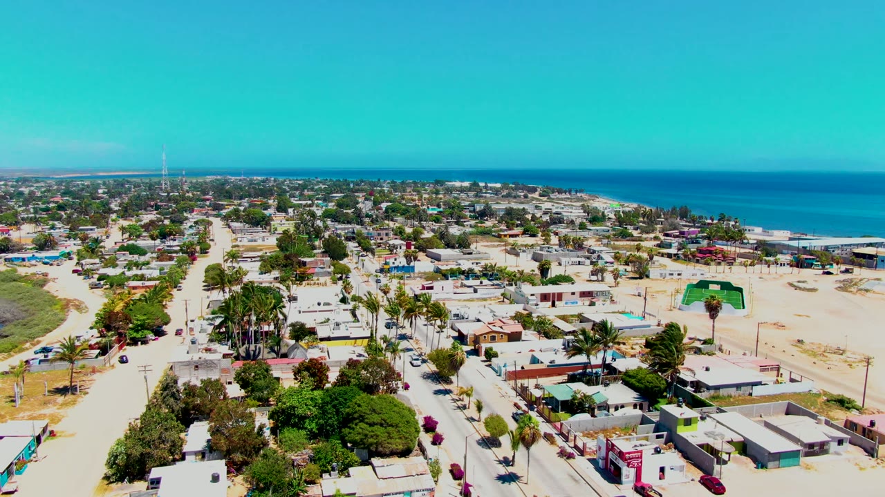 Wide aerial frame over San Carlos, Baja California Sur, showing low homes, sandy streets, palms, a blue ocean horizon, and no visible people.