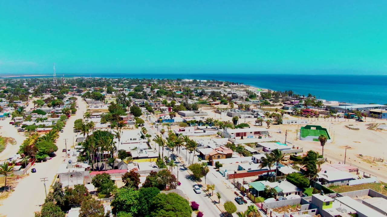 Aerial frame of San Carlos, Baja California Sur, showing a residential block with palms, parked and moving cars, sandy ground, ocean water beyond, and no visible people.