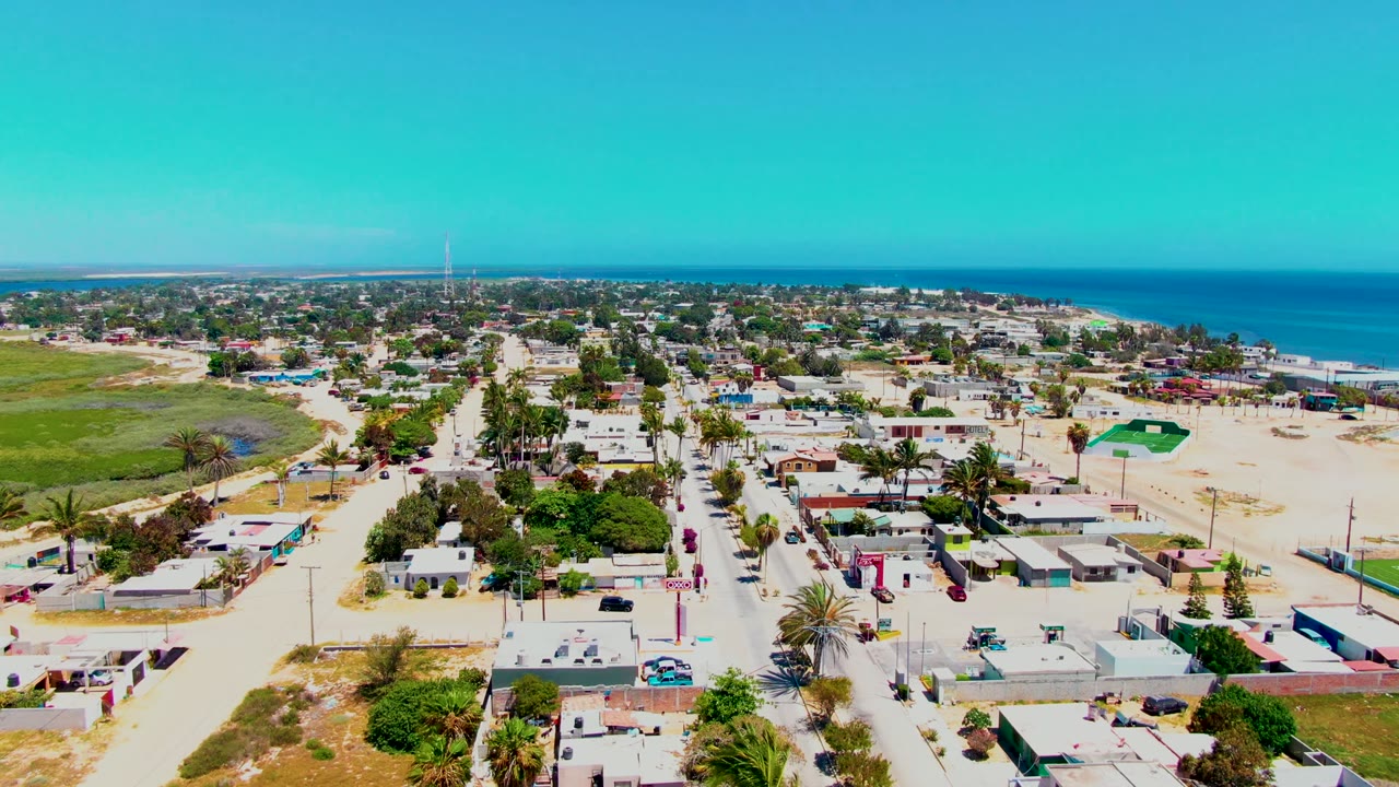 Opening aerial frame over San Carlos, Baja California Sur, showing sandy streets, low houses, palm trees, the beach, blue ocean, and no visible people.
