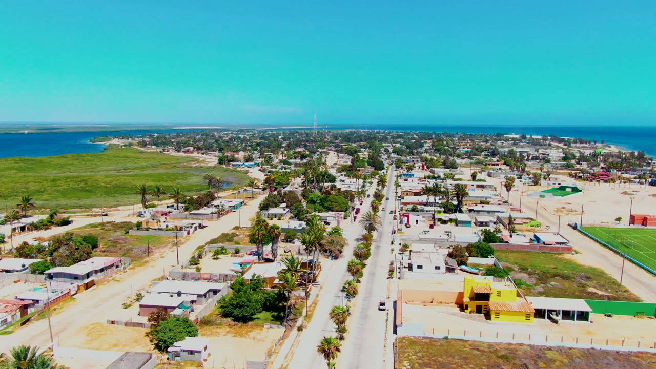 Opening aerial frame over San Carlos, Baja California Sur, with a straight road, low homes, sandy lots, palms, green field, blue ocean, and no visible people.