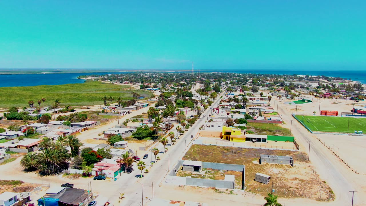Opening aerial frame over San Carlos, Baja California Sur, showing a straight residential road, low houses, sandy lots, palms, green fields, blue ocean, and no visible people.