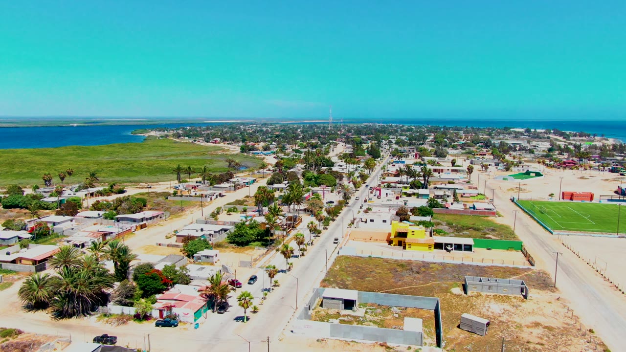 Wide drone view of road traffic through a sandy coastal neighborhood beside green fields and ocean.