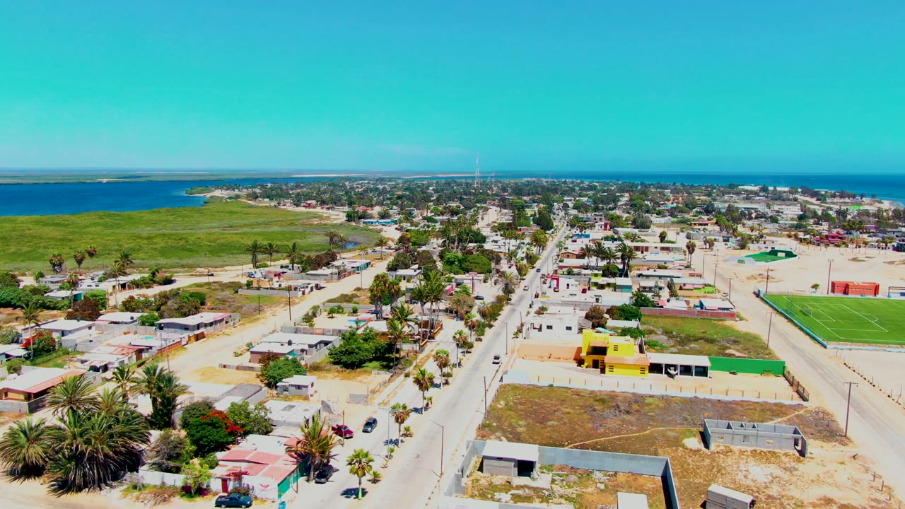 Mid-clip aerial view over San Carlos, Baja California Sur, keeping the straight road centered between low homes, palms, a green field, distant water, and no visible people.