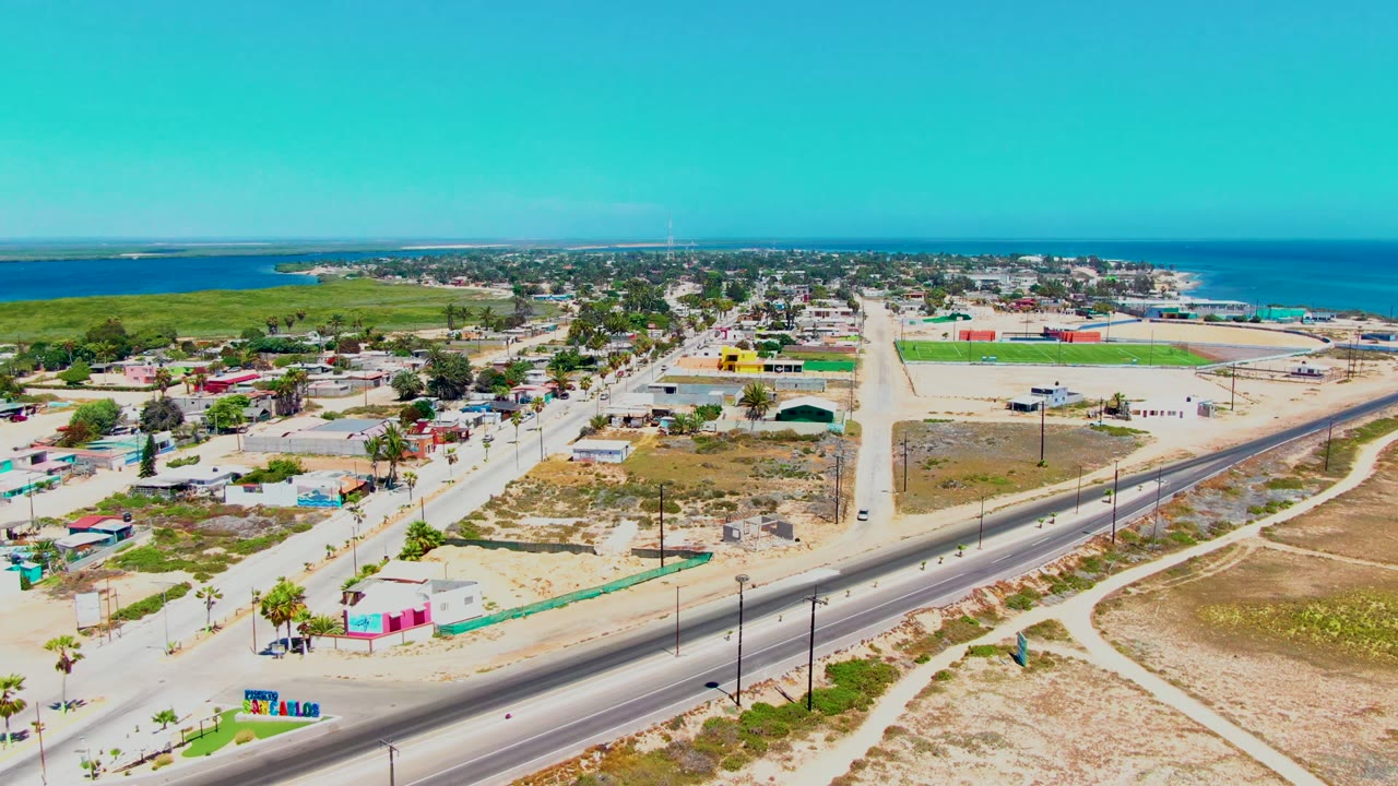 Opening aerial frame over San Carlos, Baja California Sur, showing a paved road, sandy open lots, low buildings, green fields near the water, blue ocean on the horizon, and no visible people.