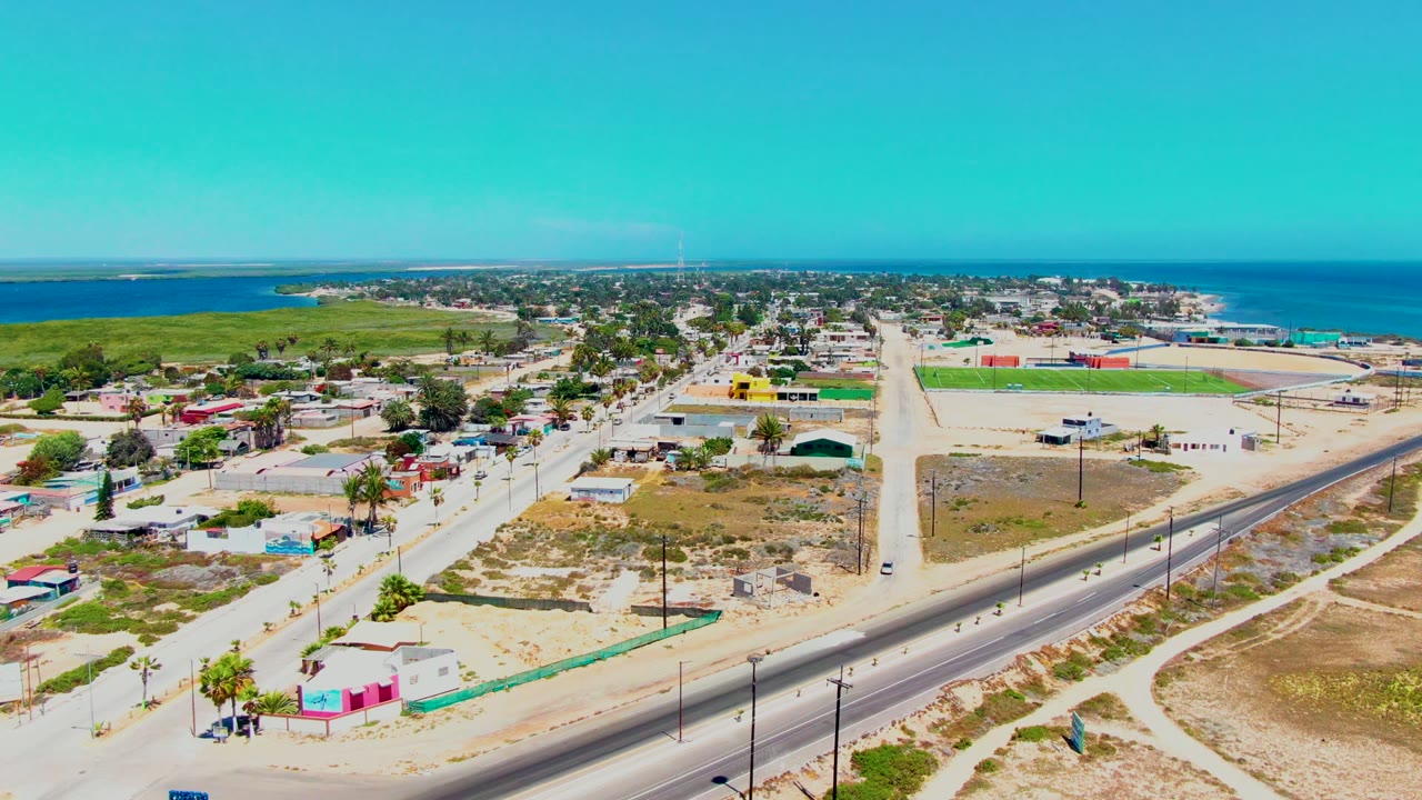 Drone view over a coastal town with a main road, buildings, sandy ground, and blue water beyond.