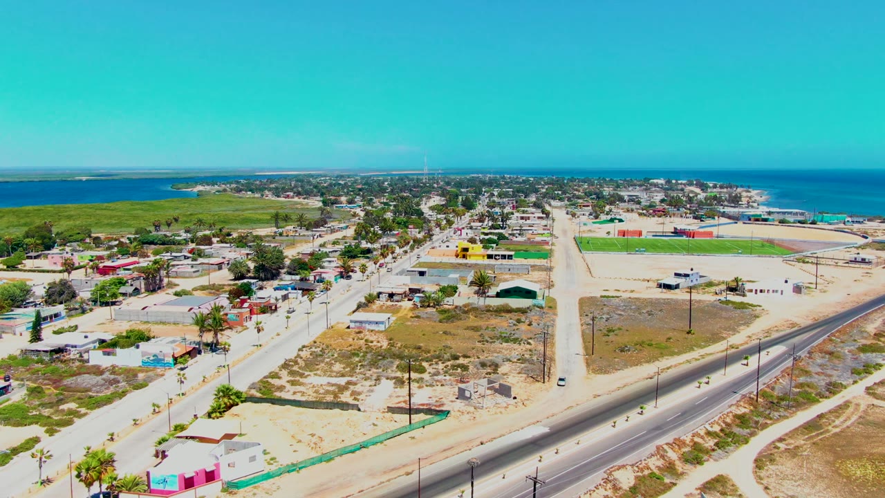 Mid-clip aerial frame in San Carlos, Baja California Sur, showing dry lots and roads in the foreground, compact town blocks and a bright green field near the shore, blue ocean at the back, and no visible people.