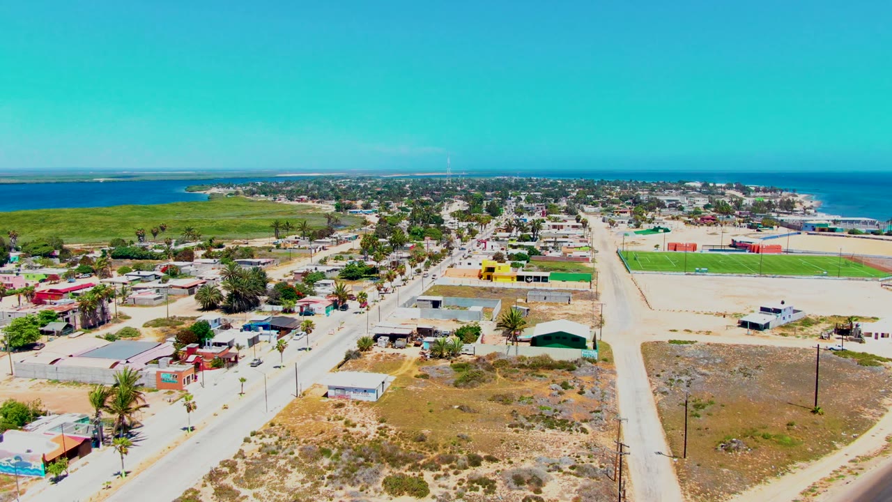 Opening aerial frame over San Carlos, Baja California Sur with sandy roads, low homes, palm trees, a green field to the right, blue ocean beyond, and no visible people.