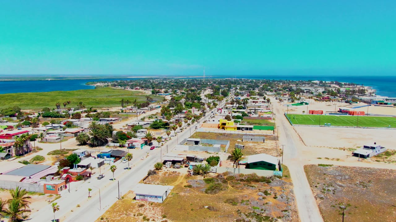 San Carlos aerial view with a central sandy road, residential rooftops, a bright green rectangular field, dry ground in the foreground, ocean in the background, and no visible people.