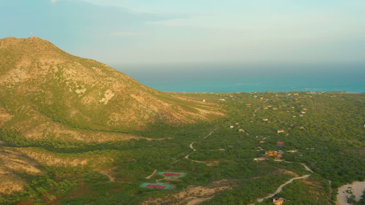 Aerial view in Cabo Pulmo, Baja California Sur, with a rocky sunlit mountain at left, green forest below, dirt roads, scattered buildings, blue ocean in the background, and no visible people.