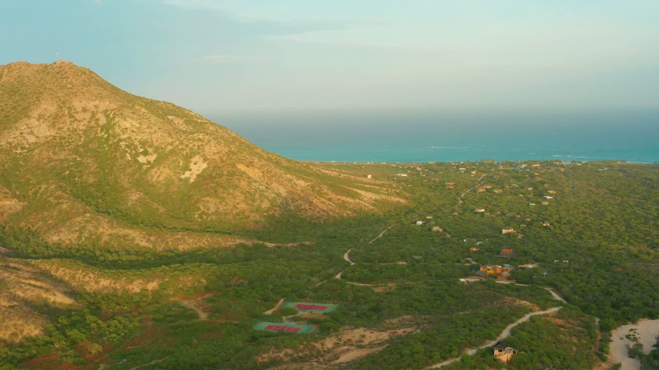 Rocky mountain above green vegetation with ocean and scattered buildings beyond.