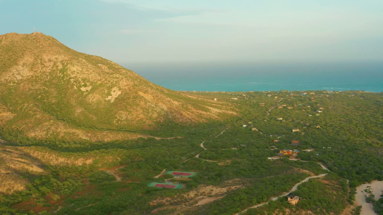 Wide aerial view over Cabo Pulmo showing a sunlit tan and green mountain, forested terrain, a pale winding road, scattered buildings, the distant blue sea, and no visible people.