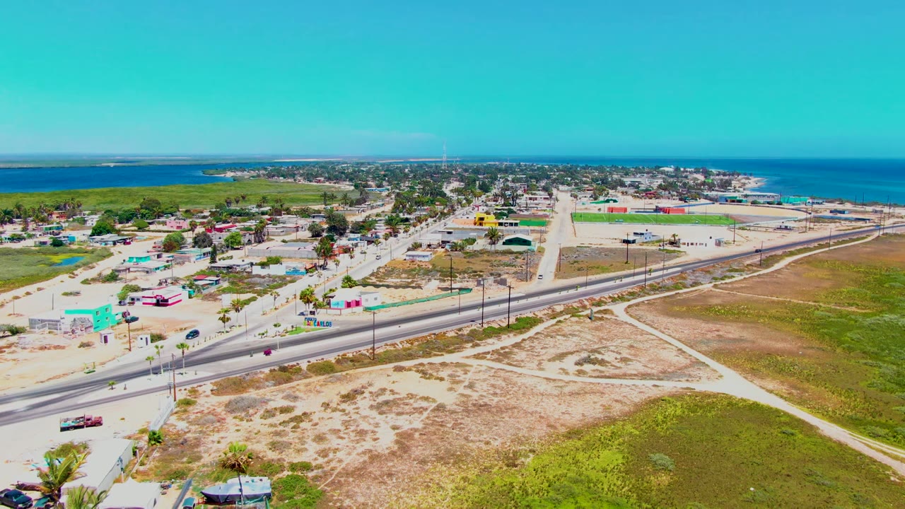 Wide aerial view over San Carlos, Baja California Sur, showing a coastal road, scattered vehicles, dry open land, small buildings, distant ocean, and no visible people.